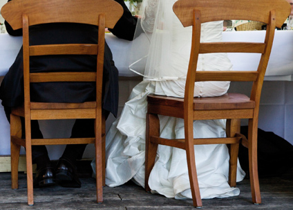 Brautpaar an Hochzeitstafel: Bräutigam im schwarzem klassischem Anzug und Braut mit üppigem Schleier und weißem Brautkleid mit vielen kleinen Köpfen von hinten zu sehen, sitzend auf antiken Holzstühlen an gedecktem festlichem Tisch